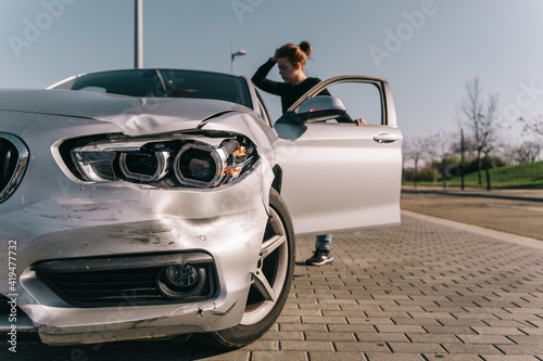 Full body of young stressed female driver in casual clothes touching head while standing on asphalt road and looking at damaged bonnet with opened mouth after accident photo