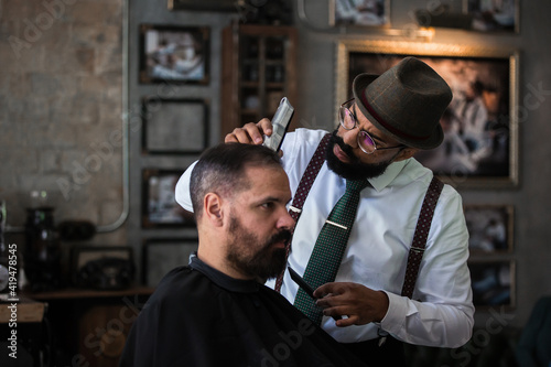Stylish dandy serious ethnic male barber trimming hair of adult client with electric clipper in hairdressing salon photo