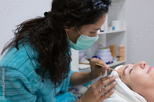 Crop anonymous cosmetologist applying hot wax with applicator on eyebrow of woman with closed eyes during epilation procedure in spa center photo