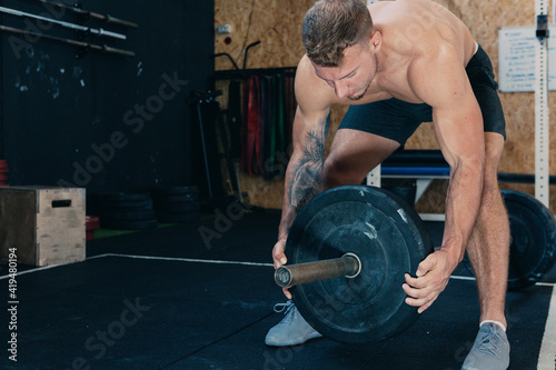 Muscular sportsman with strong naked body putting heavy weight plate on barbell while preparing for functional training in gym photo