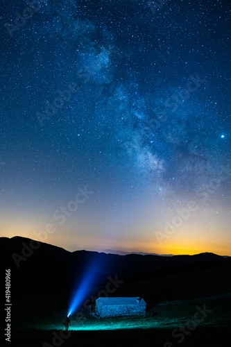Anonymous traveler silhouette with blue light standing near mountain under colorful sky with stars at night