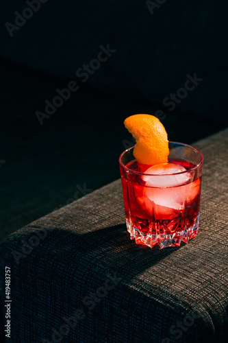 Glass of refreshing Negroni cocktail with bitter flavor and ice garnished with orange peel and served on couch arm in dark room