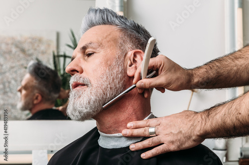 Low angle of crop unrecognizable male barber shaving gray beard of middle aged stylish client in eyeglasses sitting near mirror in hairdressing salon