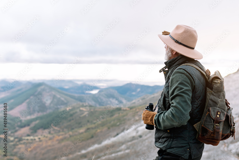 Senior male traveler in outerwear standing with binoculars in highland terrain and admiring picturesque landscape during winter holidays in Caceres Senior male traveler in outerwear standing with binoculars in highland terrain and admiring picturesque landscape during winter holidays in Caceres