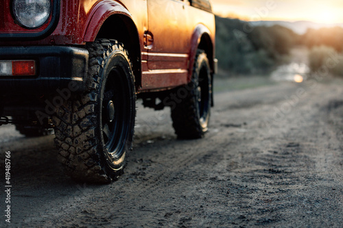 Cropped modern red off road car parked on dusty dirt road leading through forest in summer evening in countryside