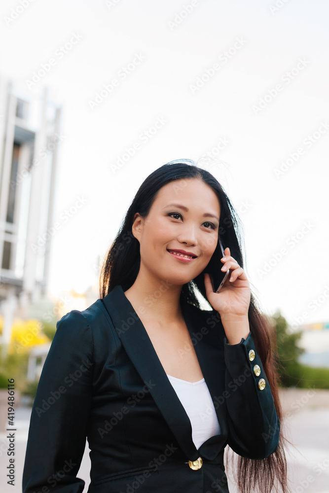 Happy Asian female entrepreneur in smart casual style standing in street of downtown browsing on mobile phone while looking away Happy Asian female entrepreneur in smart casual style standing in street of downtown browsing on mobile phone while looking away