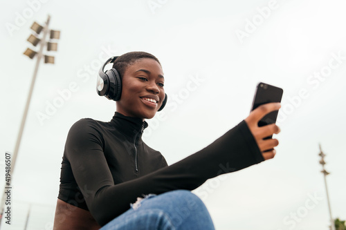 Low angle of young glad ethnic female in wireless headphones surfing internet on cellphone while listening to song in city photo
