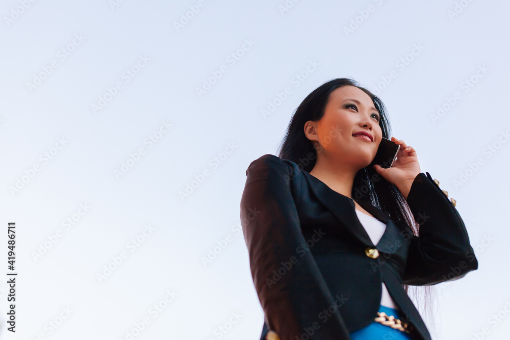 From below of Asian female entrepreneur in smart casual style standing in street of downtown and talking on mobile phone while looking away From below of Asian female entrepreneur in smart casual style standing in street of downtown and talking on mobile phone while looking away