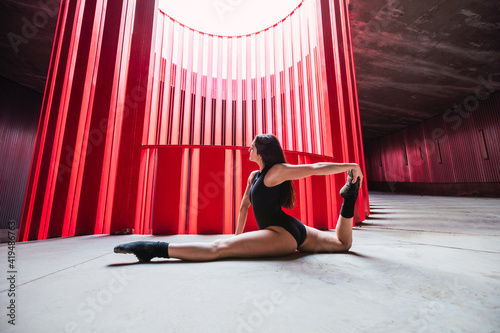 Side view of flexible female dancer sitting in split while leaning on hand in bright auditorium in daylight