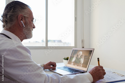 Side view of elderly male doctor in wireless earbud speaking with woman on video call on netbook in clinic