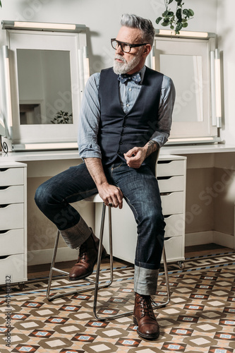 Trendy mature hipster businessman in eyewear and trendy clothes looking away on chair in house photo