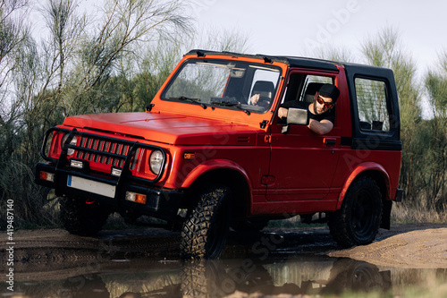 Unrecognizable man driving red SUV vehicle on dirt road with water puddle among green trees in summer countryside photo