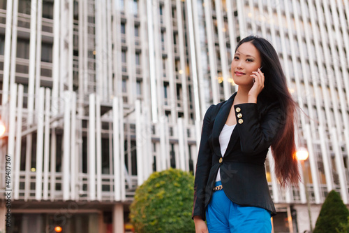 From below of Asian female entrepreneur in smart casual style standing in street of downtown and talking on mobile phone while looking away photo