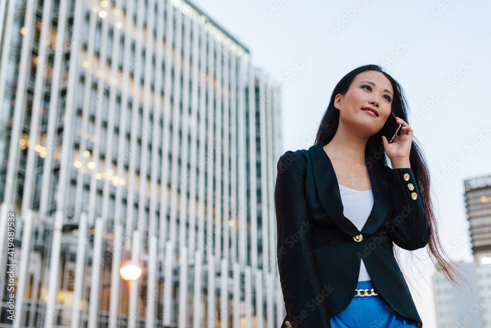Low angle of Asian female entrepreneur standing in street of megalopolis and speaking on mobile phone while discussing business issues and looking away Low angle of Asian female entrepreneur standing in street of megalopolis and speaking on mobile phone while discussing business issues and looking away