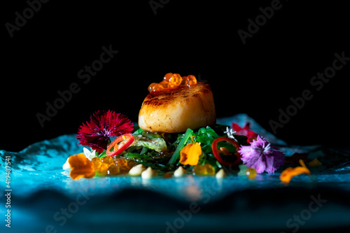 Delicious haute cuisine dish with green noodles and scallop decorated with caviar and flowers petals and served on blue surface