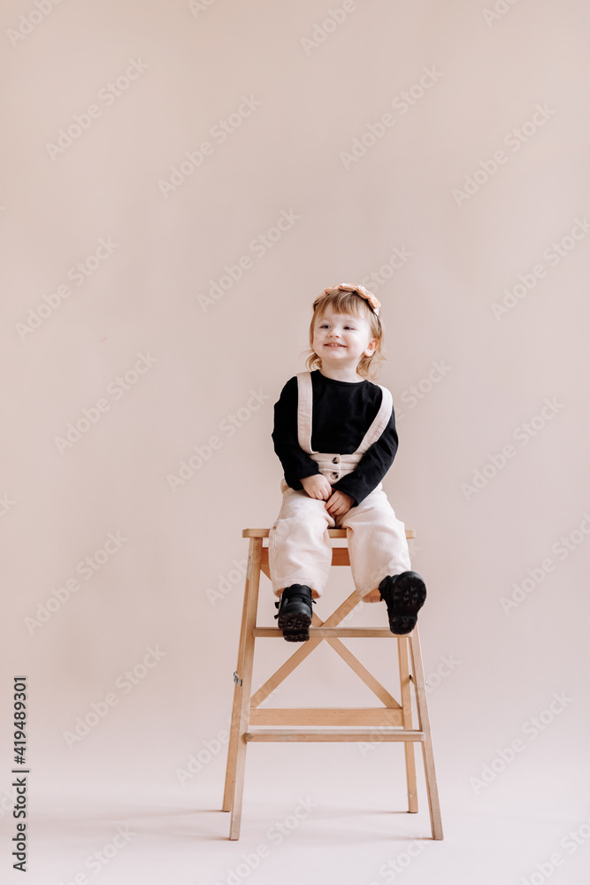 Fototapeta premium smiling little baby girl in beige jumpsuit and flower wreath on her head standing on a ladder in the studio. cute child isolated on beige background. Concept of the happy childhood.
