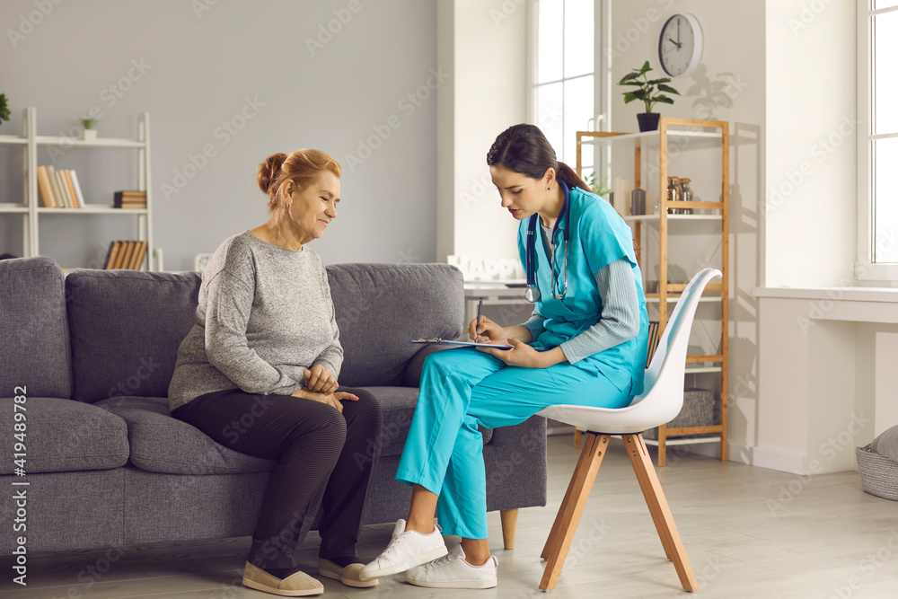 Female doctor or nurse listens to a health complaint from an elderly woman and records the information in a medical record. Mature woman sitting on sofa at home talking to nurse during her visit.