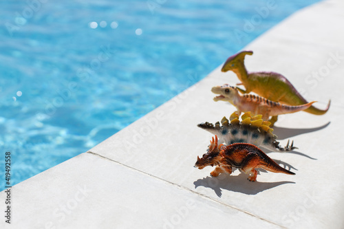 Four small toy dinosaurs stand in row near swimming pool on bright sunny day, copy space. Concept of summer vacation, childhood, togetherness, sunbathing, health. Side view. Soft focus. Horizontal