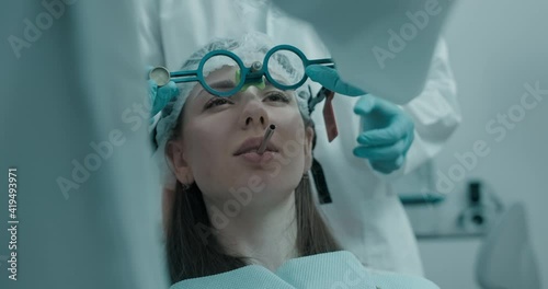 Close-up a female patient in disposable hat is sitting in the dentist chair and dentist in disposable blue gloves taking off the axiograph in front of her face.  photo