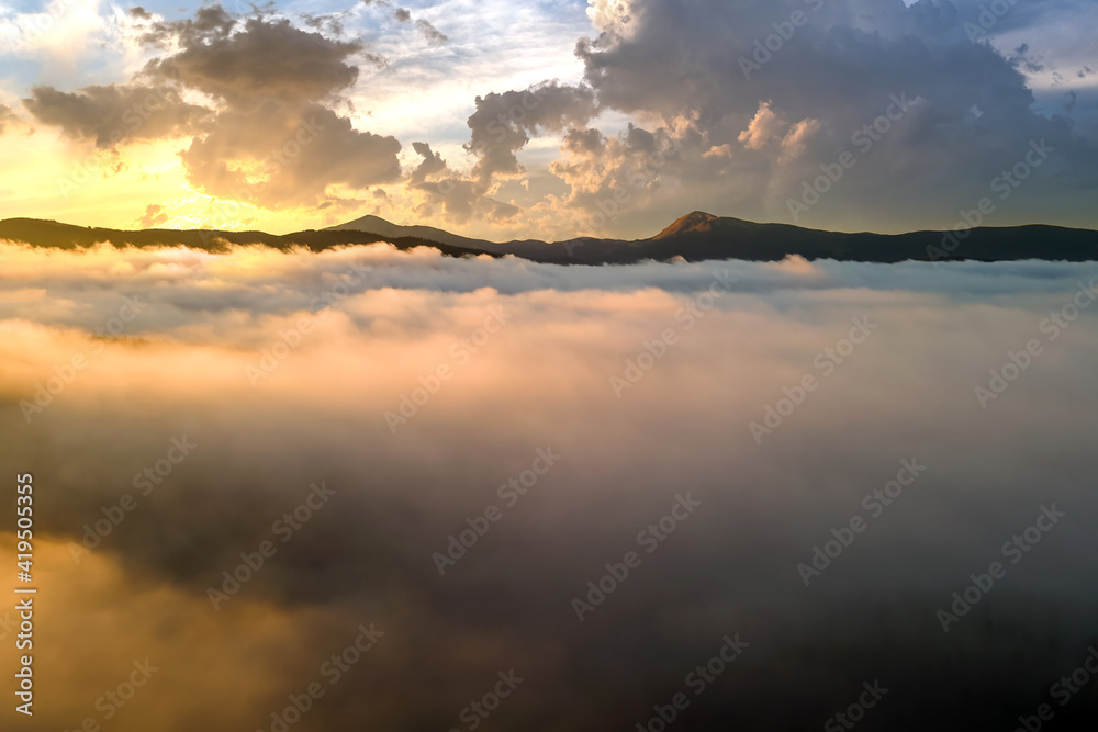 Fototapeta premium Aerial view of vibrant sunset over white dense clouds with distant dark mountains on horizon.