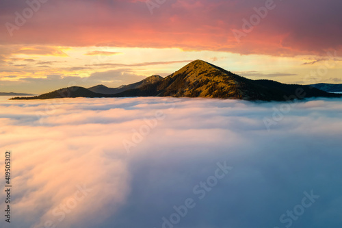 Aerial view of a big mountain over white dense clouds at bright sunrise.