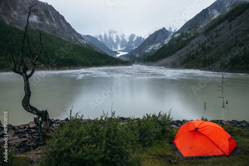 Atmospheric alpine landscape with orange tent on shore of green mountain lake and snowy mountains in rainy weather. Gloomy scenery with rainy circles on water of mountain lake and low clouds in valley