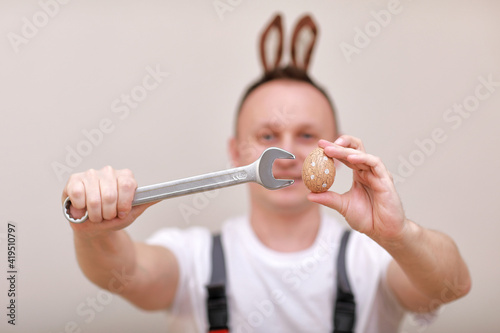 Easter holiday, celebration concept. funny smiling engineer worker or mechanic is wearing rabbit ears on head, holding wrenches and painted egg white background. celebrating orthodox day