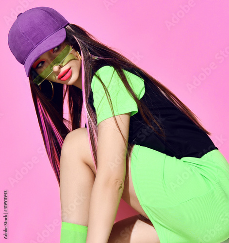 Young pretty brunette woman with purple pink colored strands in green dress, cap and sunglasses sitting and looking aside over pink background. Fashion, style, hair coloring concept