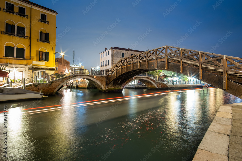 Night view of illuminated old buildings, floating boats and light reflections in canal water in Venice, Italy.