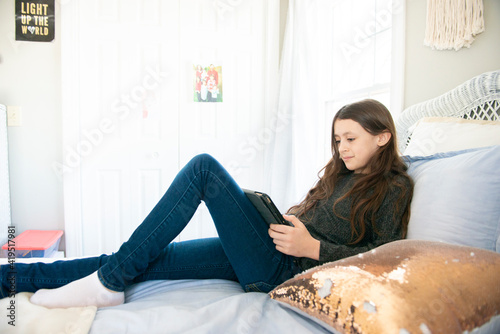 Tween girl sitting on bed with tablet in sunny bedroom.