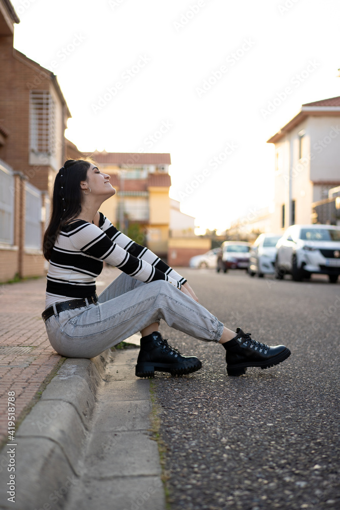 Mujer joven caucasica con camiseta de rayas negras y blancas, pelo negro, pantalón vaquero gris y botas, sentada en un bordillo junto a la carretera. Moda. Estilo