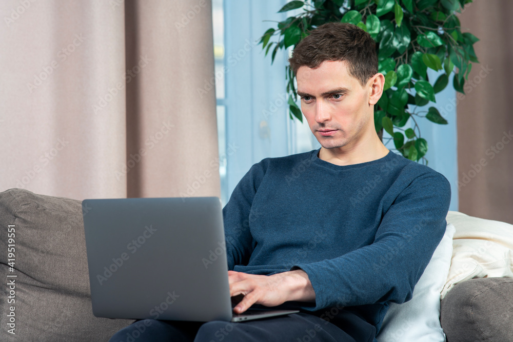 Portrait of handsome guy young man freelancer is working on his laptop computer on sofa or couch at home with a serious look, typing on keyboard, looking at screen. Freelance distant job, work concept Portrait of handsome guy young man freelancer is working on his laptop computer on sofa or couch at home with a serious look, typing on keyboard, looking at screen. Freelance distant job, work concept