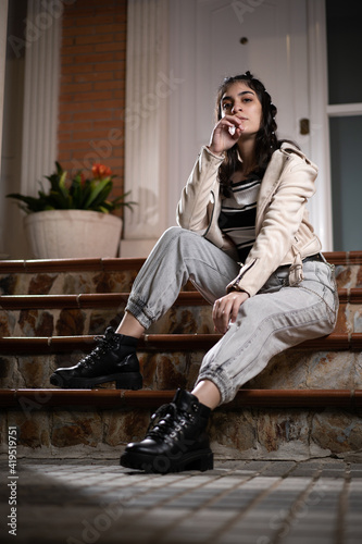 Mujer joven blanca caucásica de pelo negro y ropa urbana elegante, conc azadora blanca, camiseta de rayas blanca y negra, pantalón vaquero gris y botas, sentada posando en unas escaleras por la noche 
