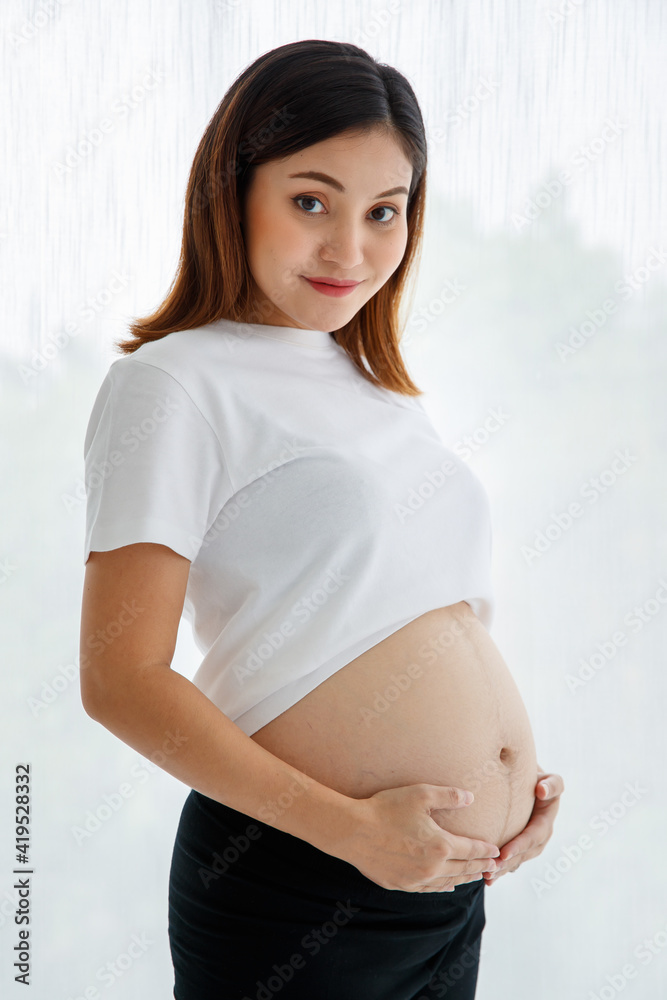 smiley young happy pregnant Asian woman wearing casually standing and touching her belly with love and care looking at camera for studio shot against white background smiley young happy pregnant Asian woman wearing casually standing and touching her belly with love and care looking at camera for studio shot against white background