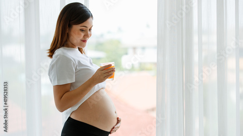 a beautiful Asian pregnant woman wearing a white T-shirt a black pants standing by the window and holding a glass of orange juice while looking at her belly with love. Family mother concept Free Download a beautiful Asian pregnant woman wearing a white T-shirt a black pants standing by the window and holding a glass of orange juice while looking at her belly with love. Family mother concept - GFXLoad