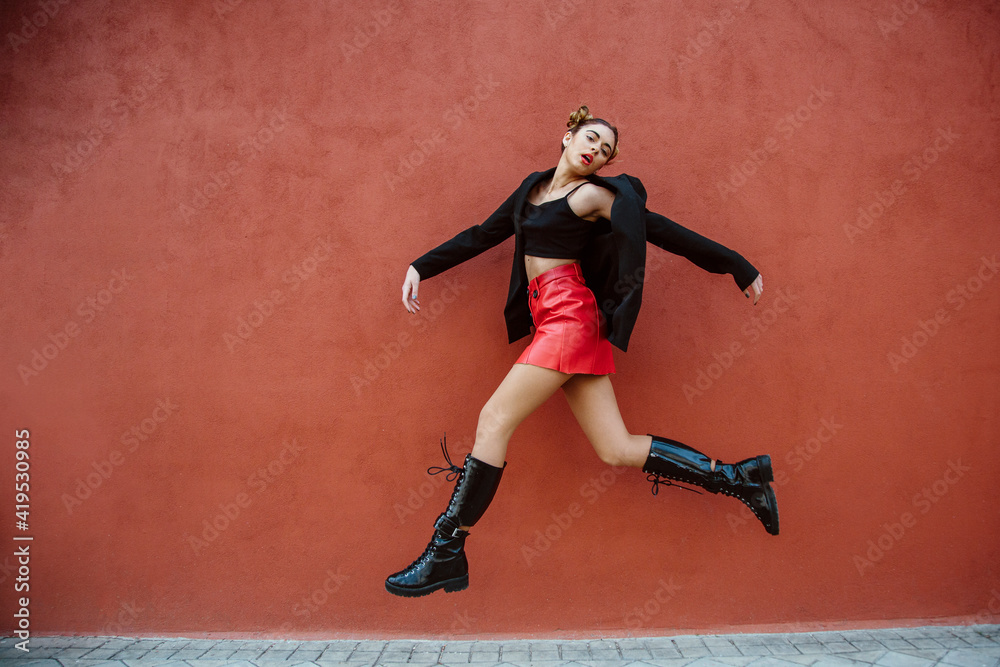 Spanish woman, with red mini skirt, jumping, looking at the camera, with a red wall of the street on the background. Spain, young women and fashion concept. Copyspace
