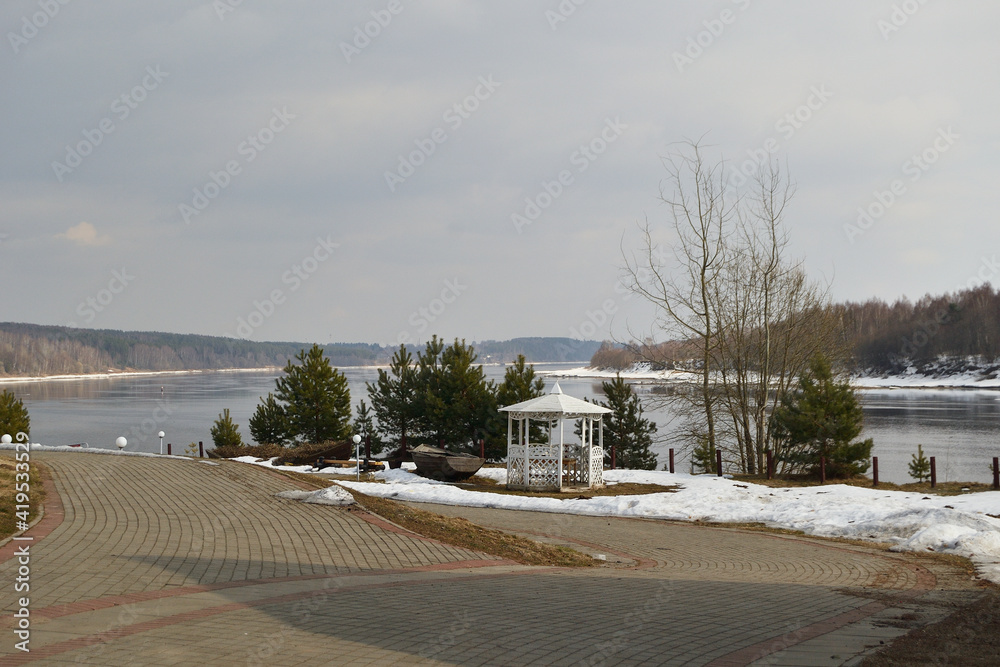 View of the white gazebo on the Volga River embankment in Demino Park, Rybinsk. Spring landscape, snow is melting, glades, cloudy dark sky, the river is being freed from ice.