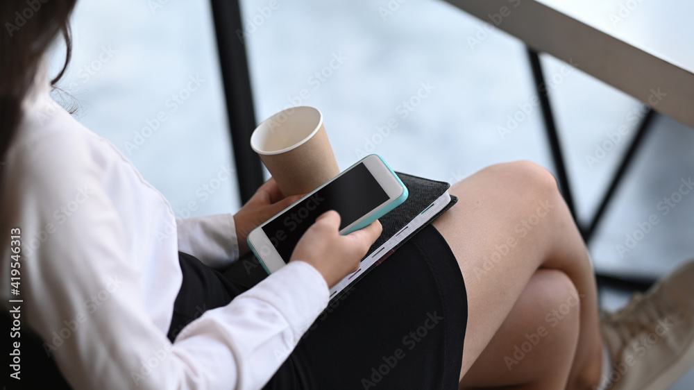 Cropped shot of casual woman holding smart phone and coffee cup while sitting in coffee shop.