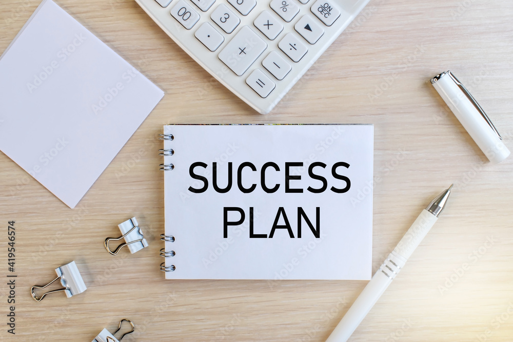 The inscription success plan on a white notebook that lies on a wooden table near a white calculator and a white pen with stationery clips. Business concept The inscription success plan on a white notebook that lies on a wooden table near a white calculator and a white pen with stationery clips. Business concept
