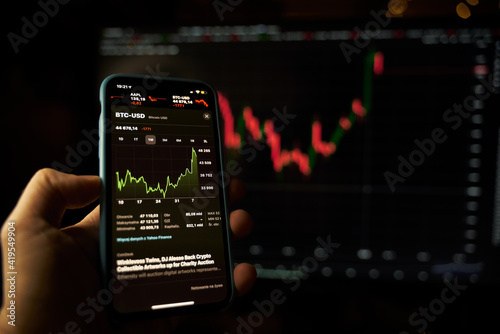 Hand holding a modern smartphone with the stocks market app and big screen blurred on the background