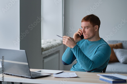 Prosperous young male freelancer making telephone call while working remotely on laptop computer in coworking office , handsome smiling hipster guy enjoying mobile phone
