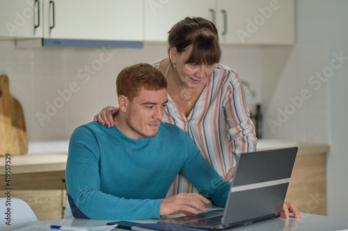 Mature woman and her adult son surfing in Internet with laptop at kitchen. Grandma shows her page on a social network. Elderly woman shares her observations online
