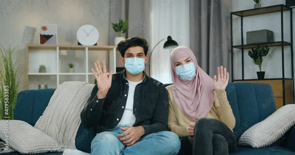 Attractive positive young arabic couple in medical masks sitting on soft couch at home during isolation of coronavirus pandemia and waving hands into camera