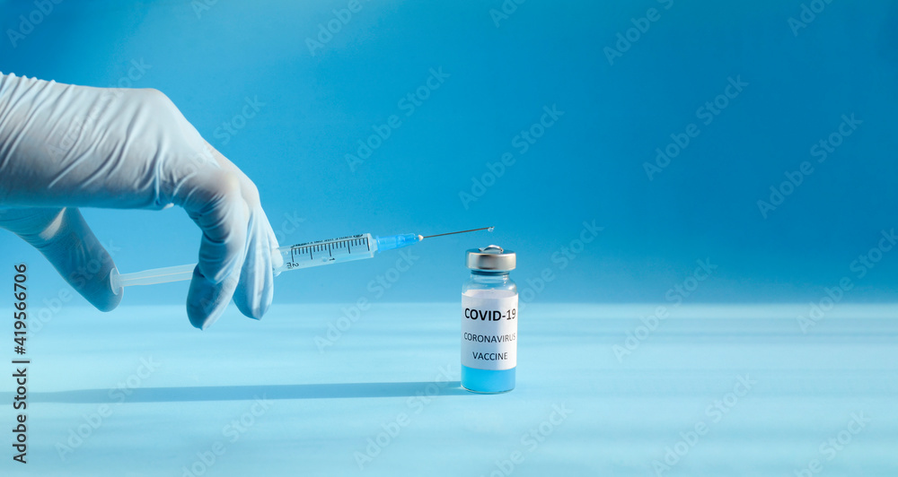 Hand in medical blue glove draws liquid into syringe for injection, Glass bottle with inscription coronavirus vaccine Covid 19 on blue background, copy space, shadows. Vaccination concept, banner .