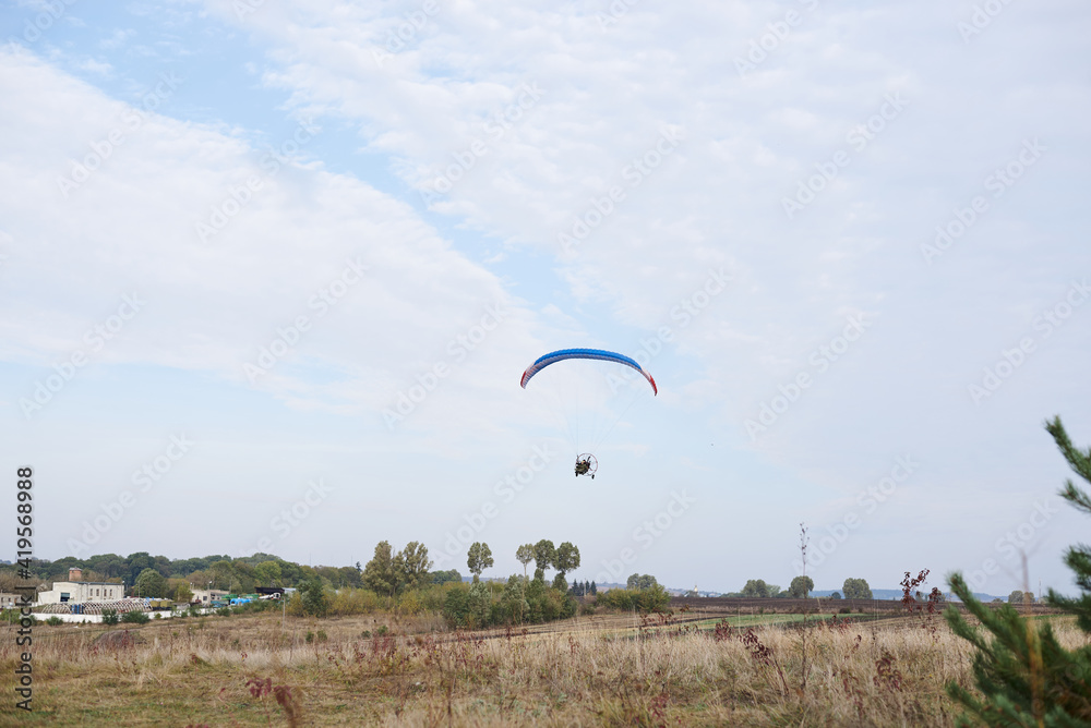 Paratrike with blue parachute flying high in the cloudy sky on autumn morning. Paragliding activity in rural countryside. Weekend leisure time and hobby. Extreme sport flight on paramotor machine. Paratrike with blue parachute flying high in the cloudy sky on autumn morning. Paragliding activity in rural countryside. Weekend leisure time and hobby. Extreme sport flight on paramotor machine.