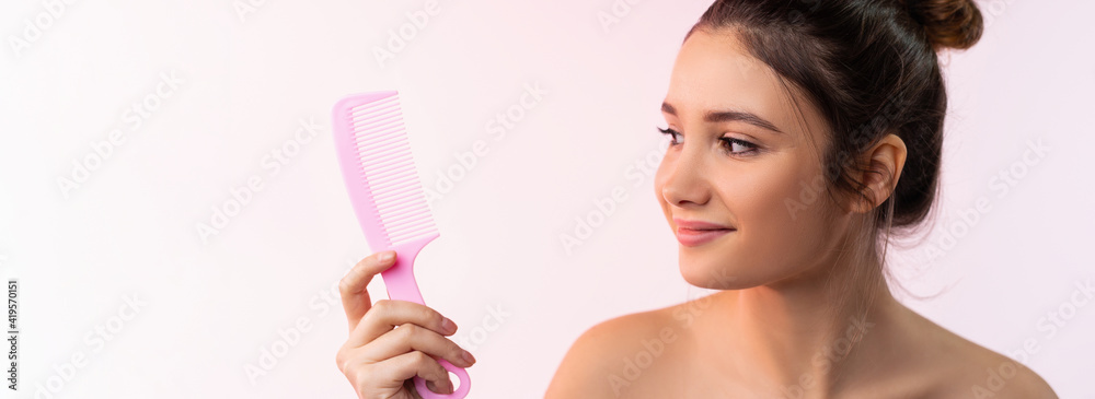Long banner in beauty and care concept. Portrait of a beautiful girl, isolated on pale pink background, with bare shoulders, holding a comb, looking at her with a smile.