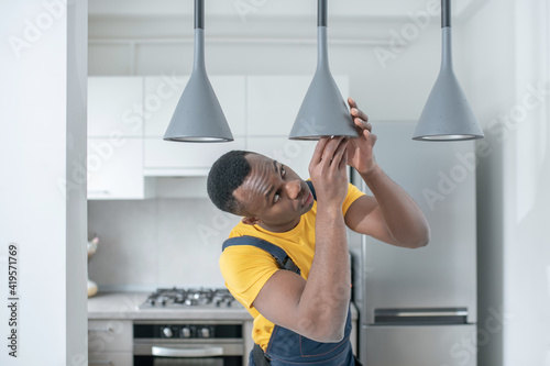 Dark-skinned service man in yellow tshirt repairing electricity lamps