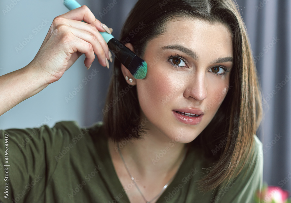 Closeup of a gorgeous brunette young woman looking directly to the camera and making makeup herself with a brush. Attractive female with natural beauty making make-up. Closeup of a gorgeous brunette young woman looking directly to the camera and making makeup herself with a brush. Attractive female with natural beauty making make-up.