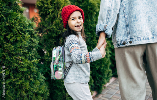 Rear view of a happy kid in a red hat going to the school with her mother by the hand. A pretty little girl with a backpack walks on the street with her mother. Mother and daughter spend time together