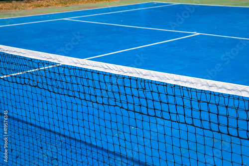 Blue tennis court with net, sport field photo. Lawn tennis on hard court. Sunny tennis court empty abstract photo with white markup and net. Tennis game play concept